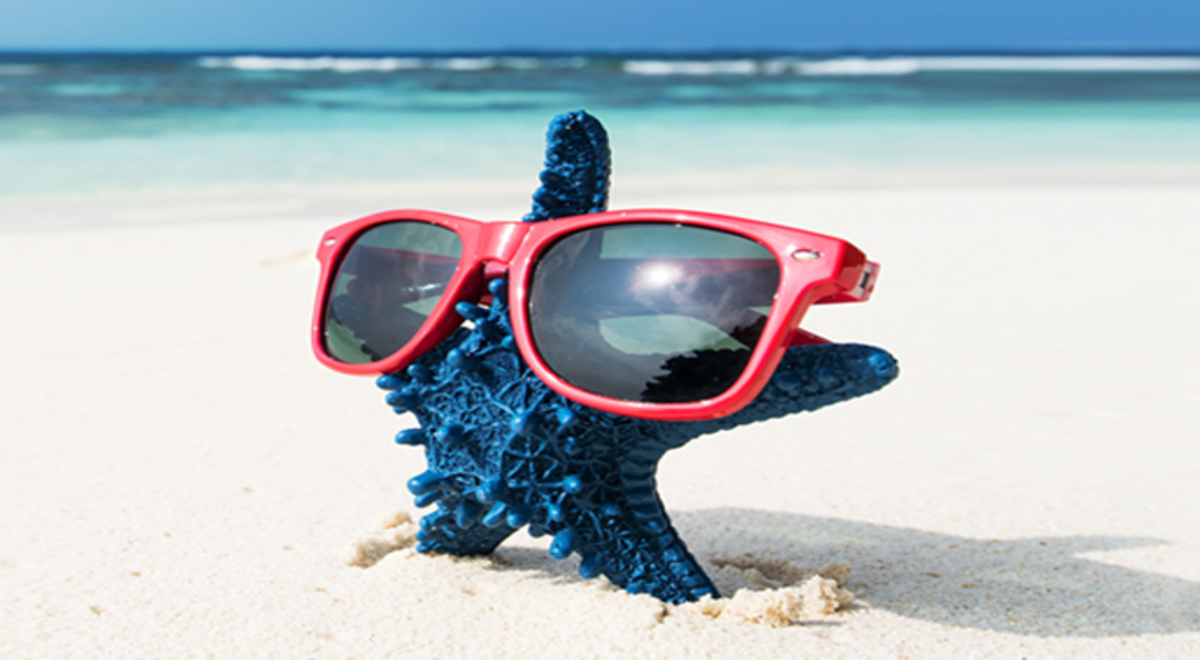 A blue starfish wears red-rimmed sunglasses as it stands on a white-sand beach with the surf in the background.