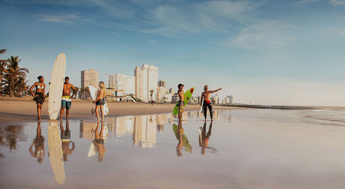 Surfies with boards stand on a broad beach looking out at the surf