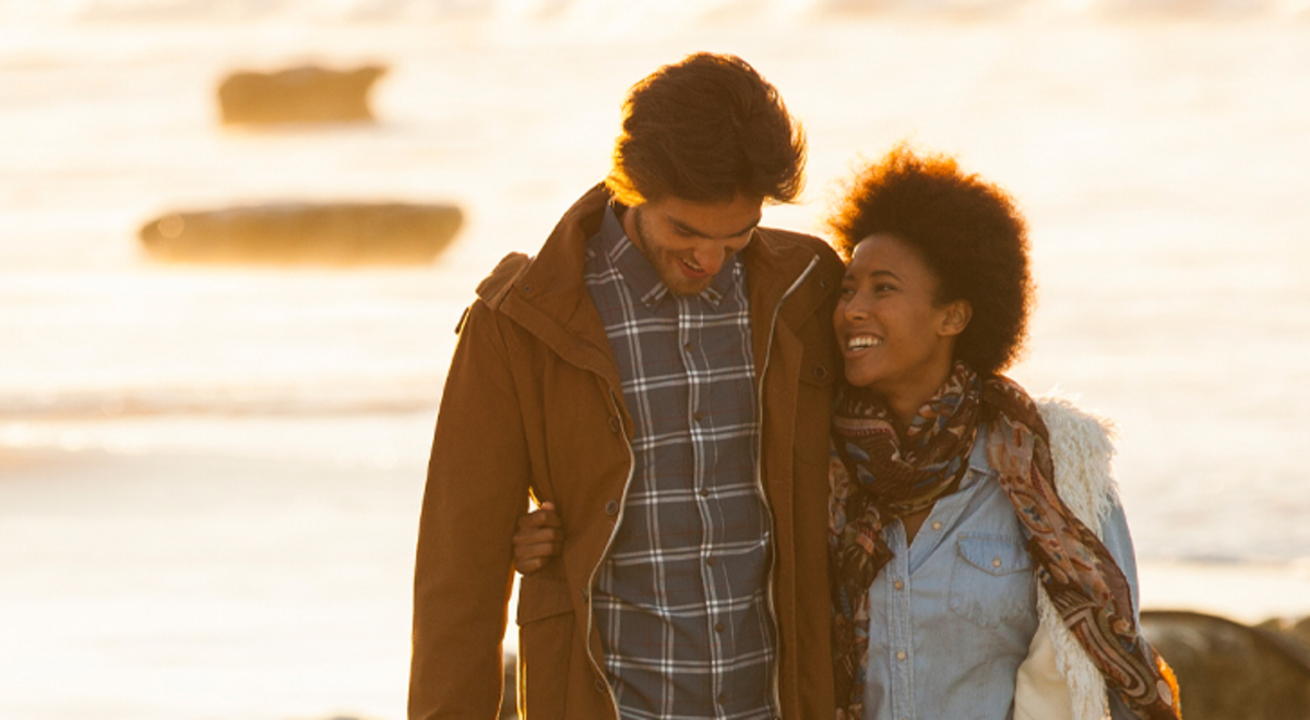 A man and women smile and walk arm-in-arm along a beach.