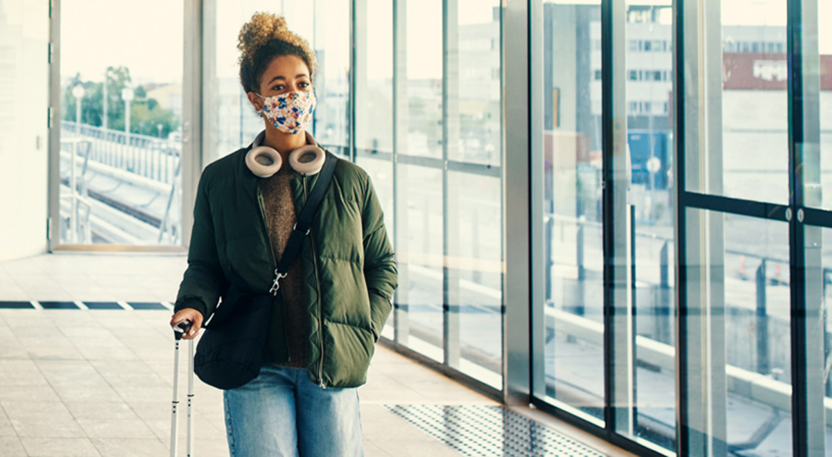 A person wearing a mask walking into an airport with a bag of wheels