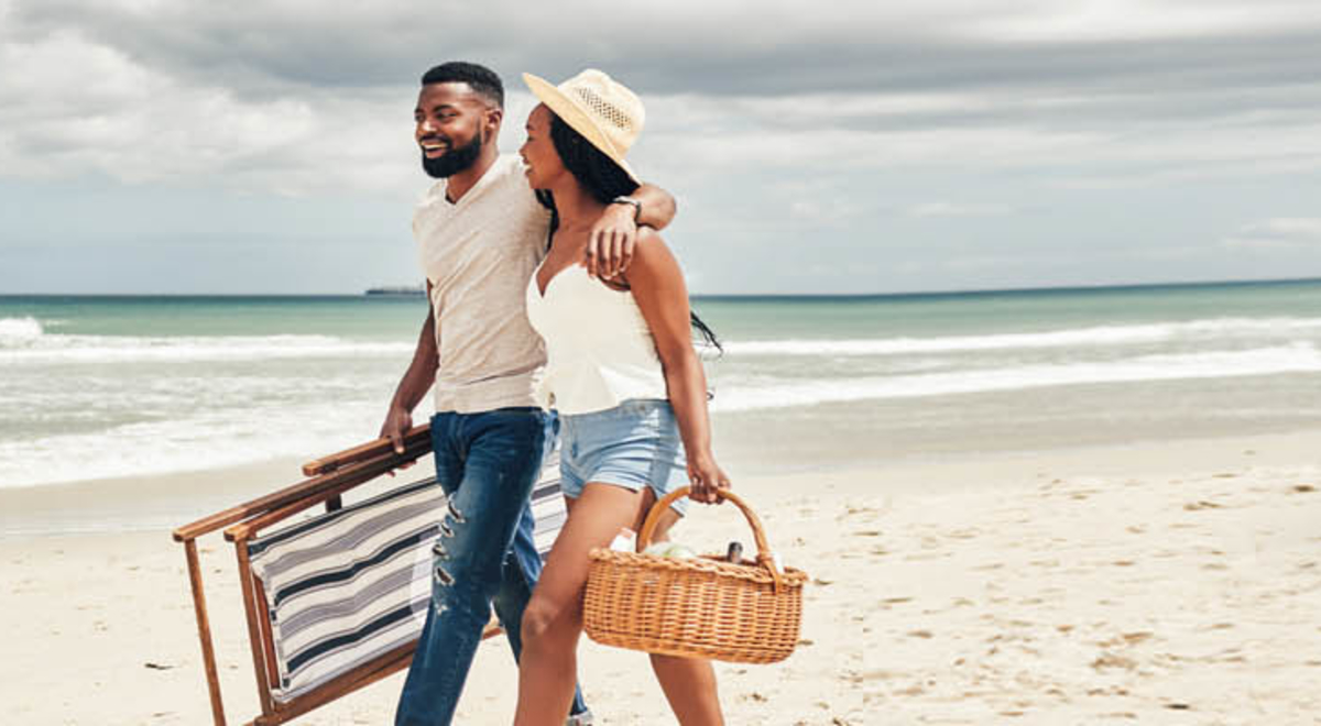 A couple hugging and walking on the beach as they hold a chair and a picnic basket