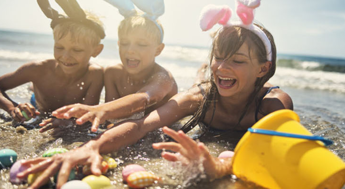 Three children wearing easter bunny ears play with buckets and spades at the edge of the shore on the beach