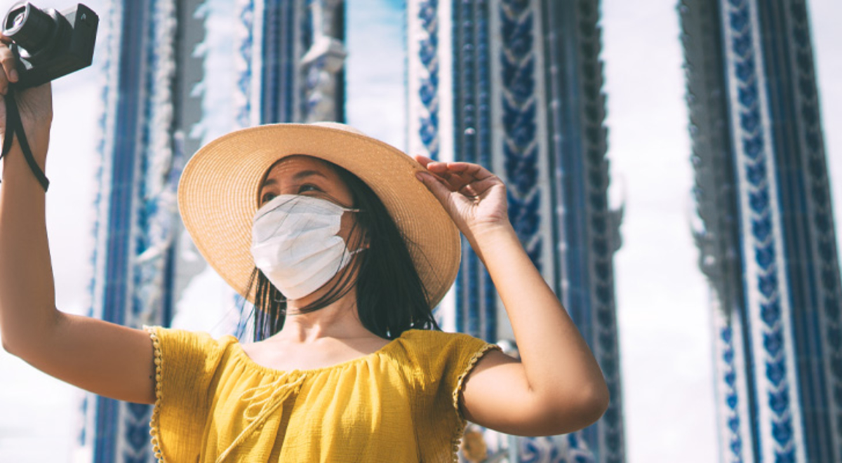 A woman in a Covid mask holds up a camera in front of a tall building.