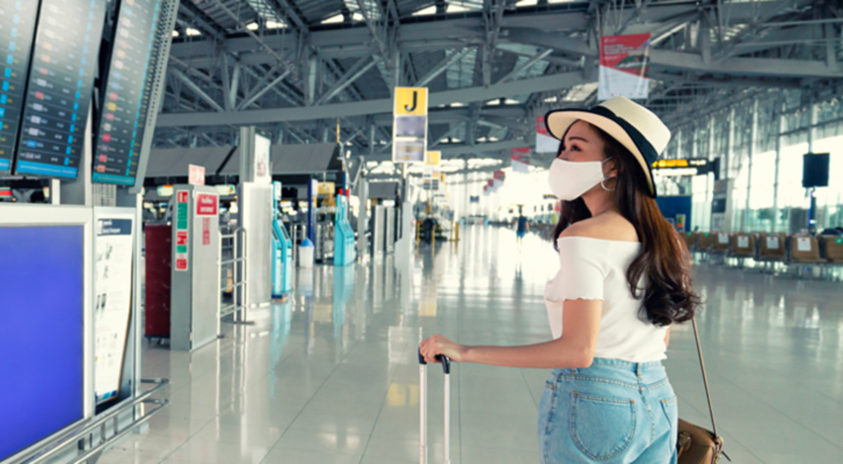 A woman with luggage wearing a covid mask looks up at digital departure boards at an airport