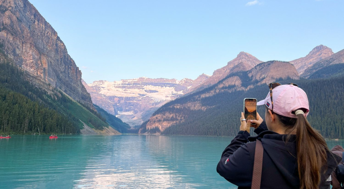 A young woman taking photos on her phone. She is taking photos of the beautiful Rocky Mountains with stunning greenery and beautiful blue waters