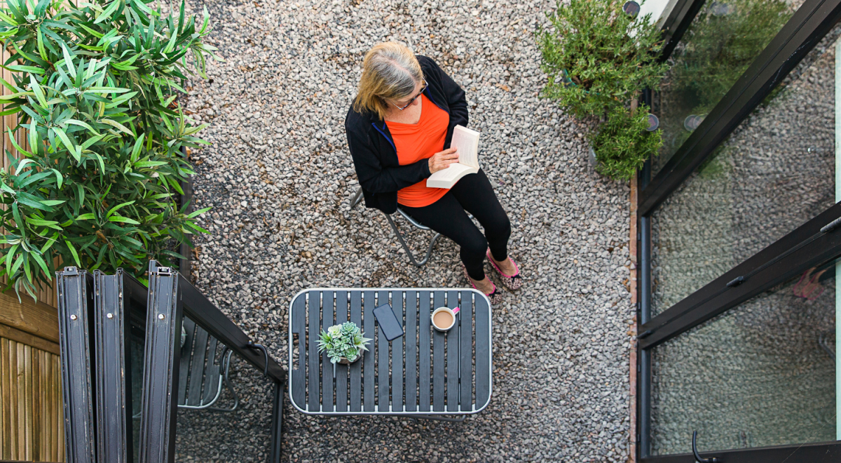 A woman in orange top reading a book while sitting on her pebbled porch