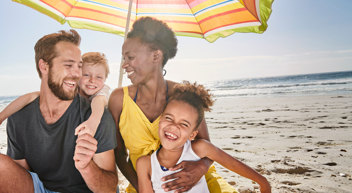 A family of four laughing and enjoying a beach vacation under a beach umbrella