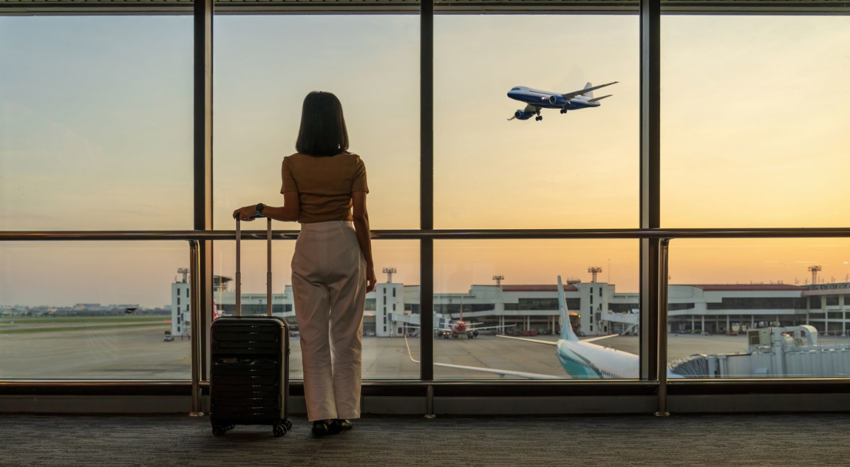 A person standing with a suitcase looking out an airport window. Through the window a plane can be seen landing.