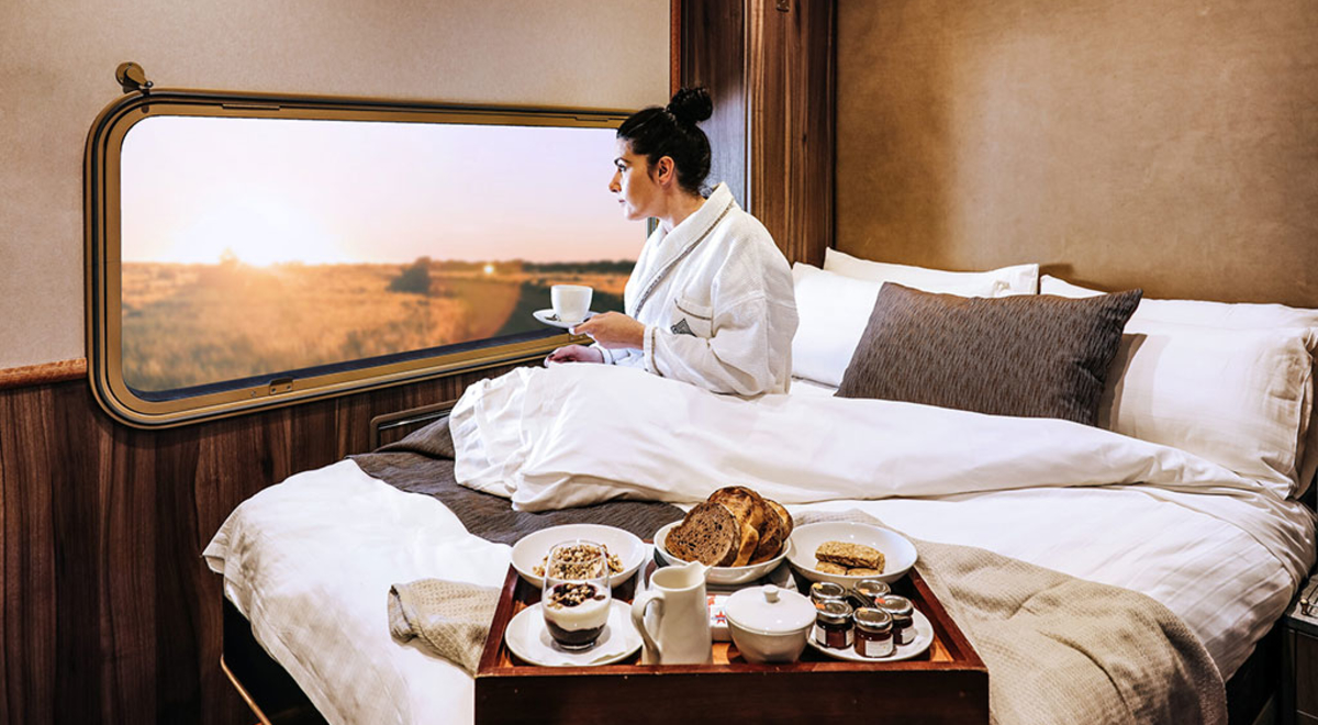Journey Beyond Platinum Service rail cabin with a woman sitting on a king-sized bed while sipping her coffee