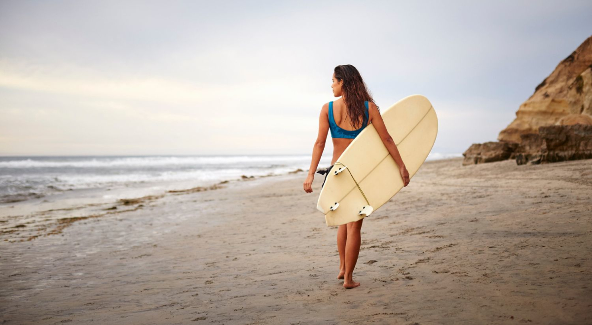 A young woman walking along a beach holding a surf board as she looks at the waves