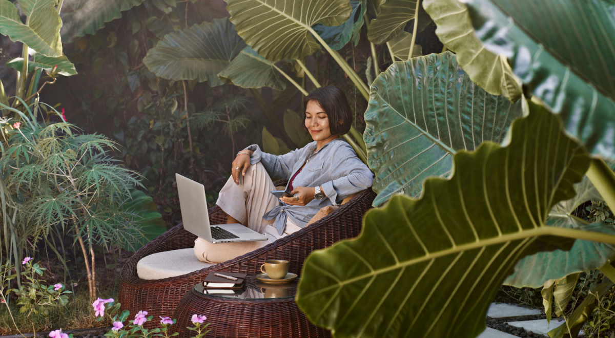person sits in tropical garden on cane chair with laptop, diary and coffee
