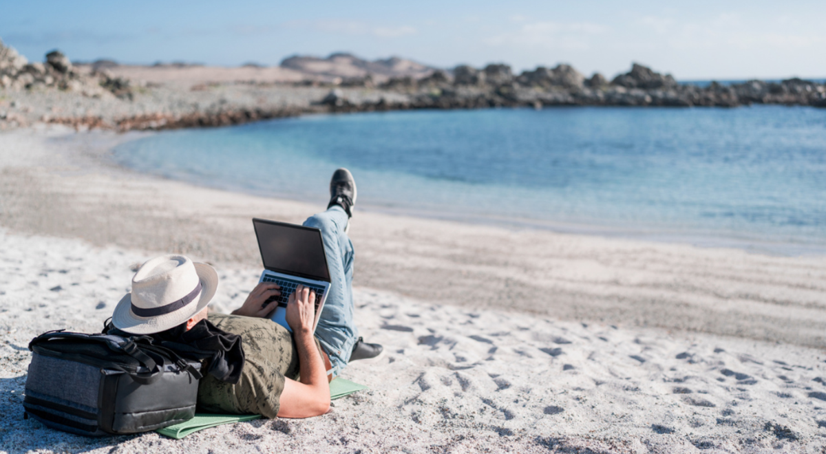 Man lying on beach resting head on backpack while working on his laptop