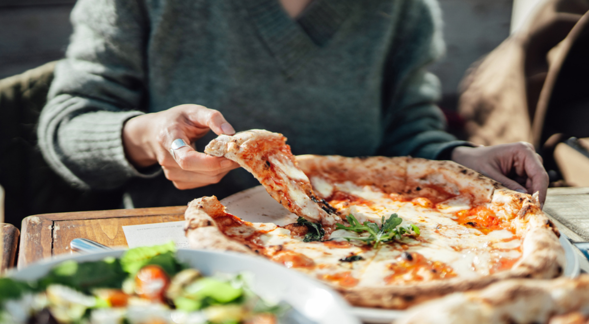 Lady wearing grey sweater lifting the first piece of pizza from board on table at restaurant