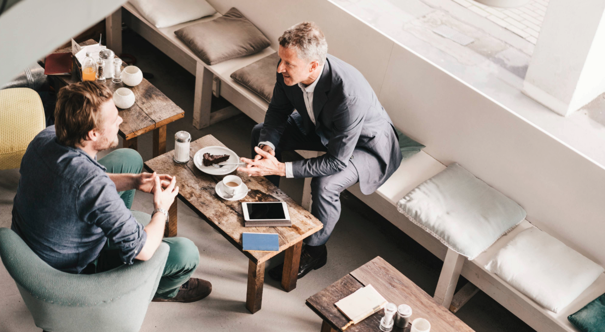 two business men sitting across from each other with cakes and coffee on small table between them