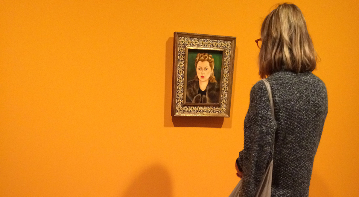 a short-haired woman staring on a portrait hung on an orange wall while a kid is playing beside her on the floor