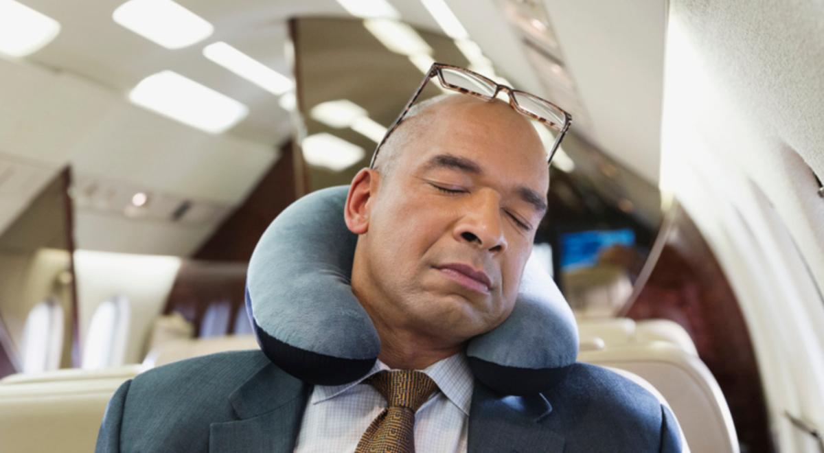 A man wearing a suite sleeping on a plane with a neck pillow around his neck