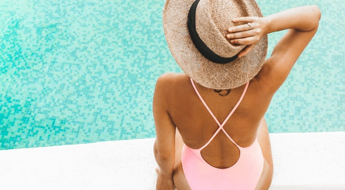 a woman on a light pink one piece bikini sitting beside the pool while holding on to her straw hat