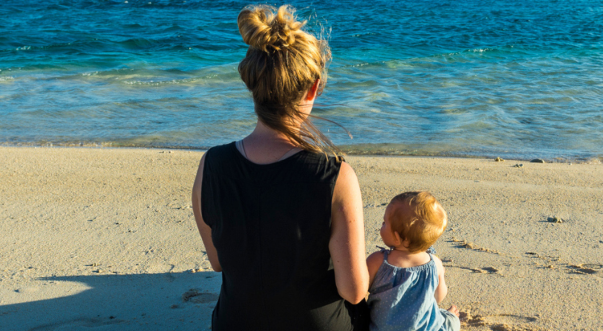 A woman and baby sit looking out to sea from a beach in Rarotonga