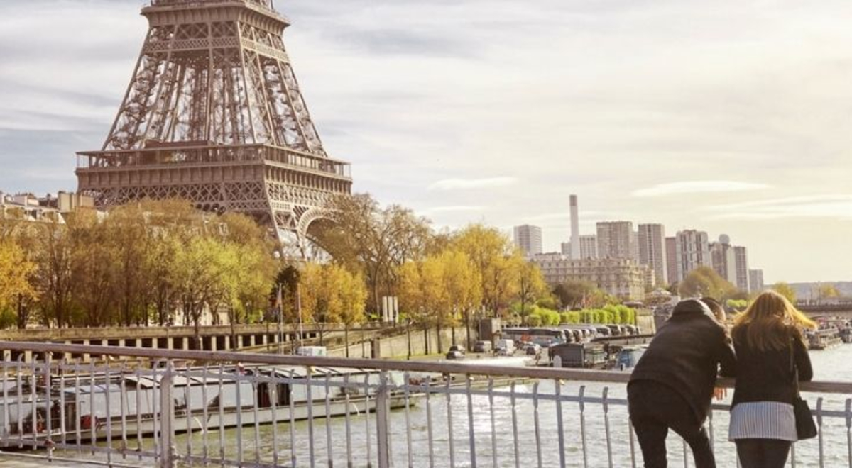 Femme and masculine presenting people stand on bridge over looking Seine River in Paris. the Eiffell Tower is in the background. 