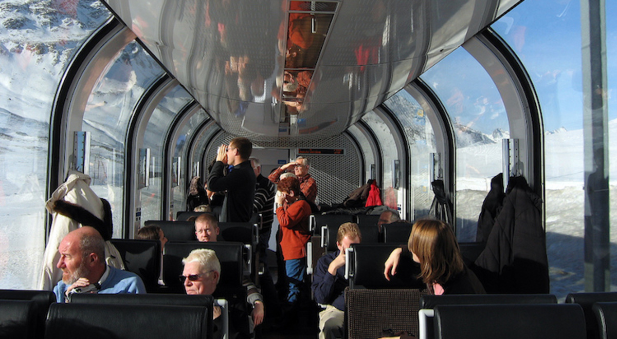 Passengers in a train carriage with high, curving viewing windows look out on a snowy, mountainous landscape as the sun pours in.