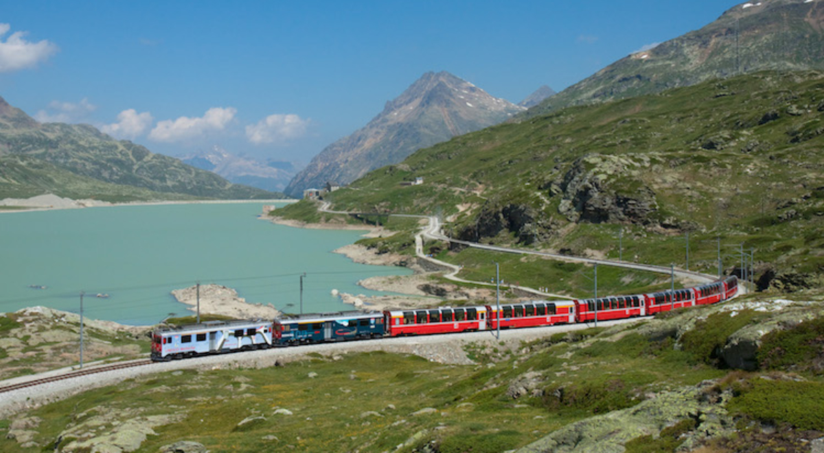 A passenger train winds its way around a lake in a green mountainous region of Switzerland