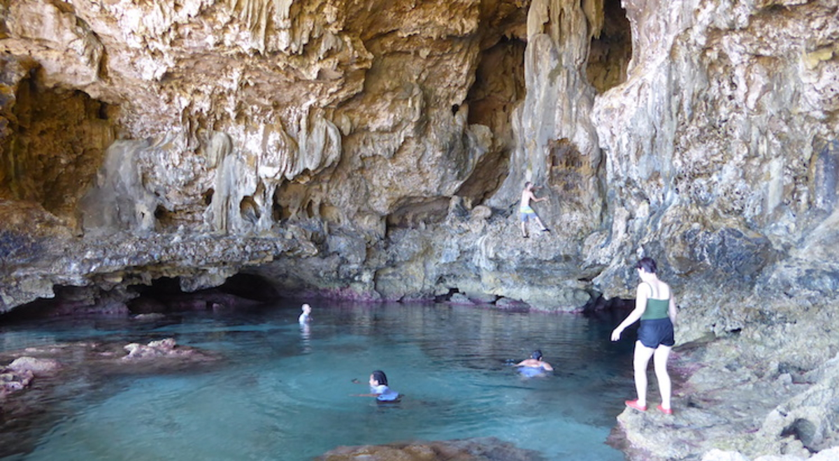 People swim in a large ocean pool in a limestone cavern with high pocked walls where other people can be seen climbing.