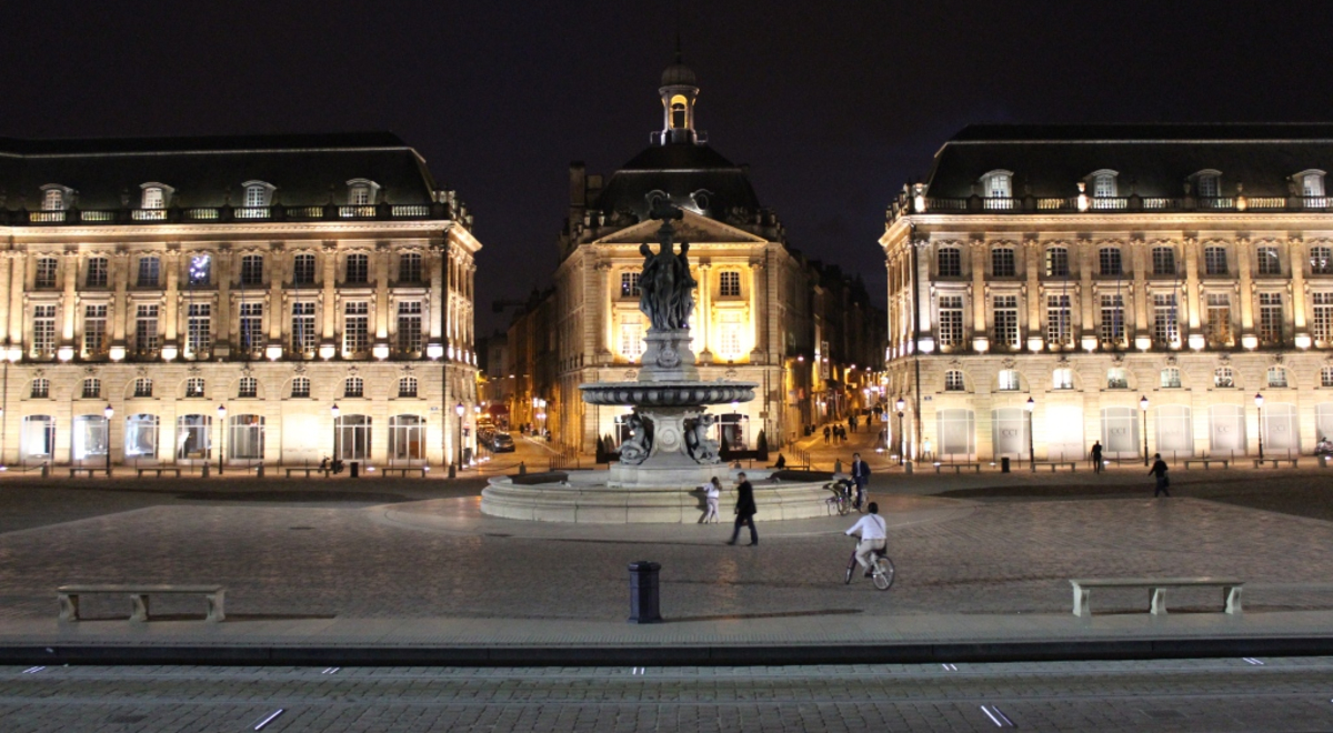 People mill around a large ornate fountain in a city square at night