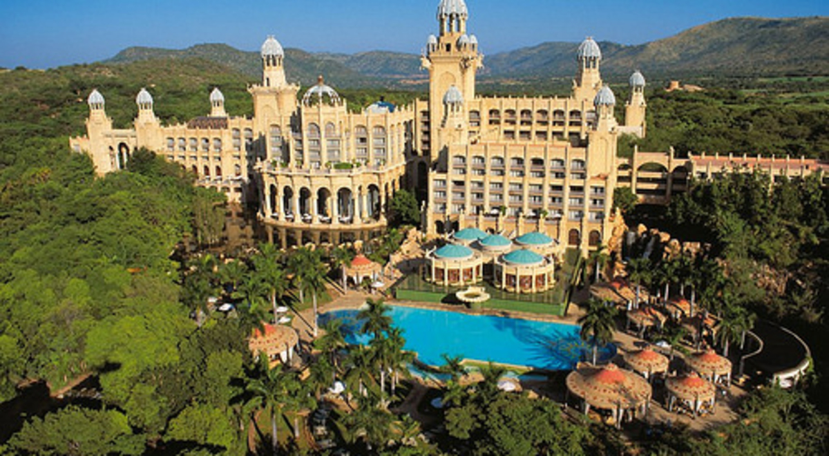 A large ornate hotel with archways and domes and a large swimming pool stands in a forest and palm trees.