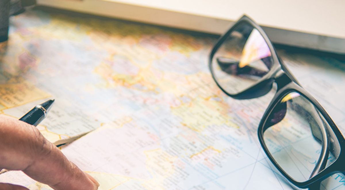 A man's hand holding a pen, a camera, sunglasses and a summer hat rest on an open atlas