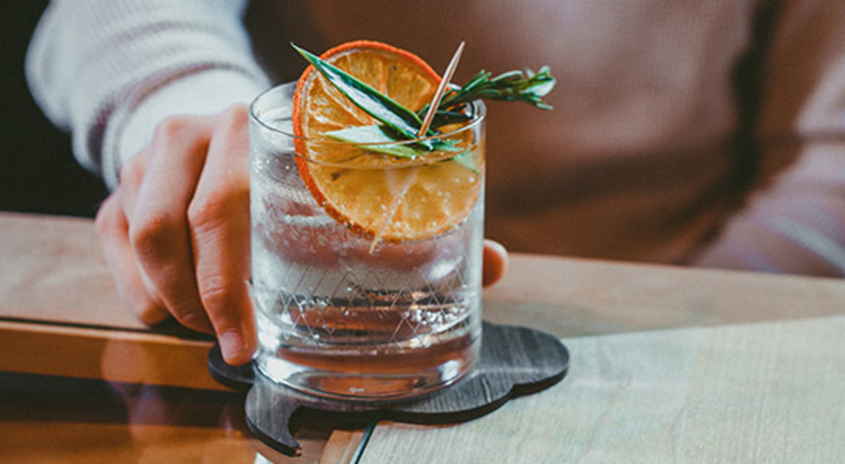 Man in grey jumper holding a glass of gin garnished with an orange slice and herbs 