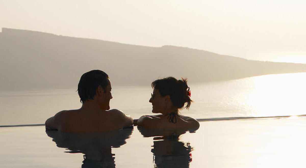 A couple swim at the edge of an infinity pool as the sun sets over distant hills and the sea.