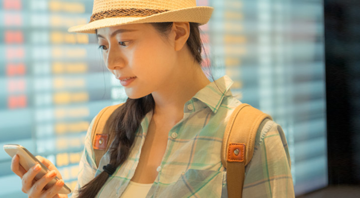 female presenting person wearing straw hat standing in front of departure board and looking at phone 