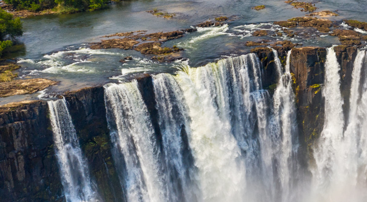 A drone's view of Victoria Falls plummeting down broad rock faces creating spray and a rainbow.