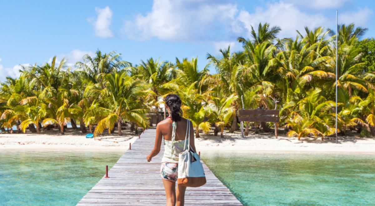 A woman walks along a jetty over calm tropical waters leading to a white-sand beach and a forest of palm trees.