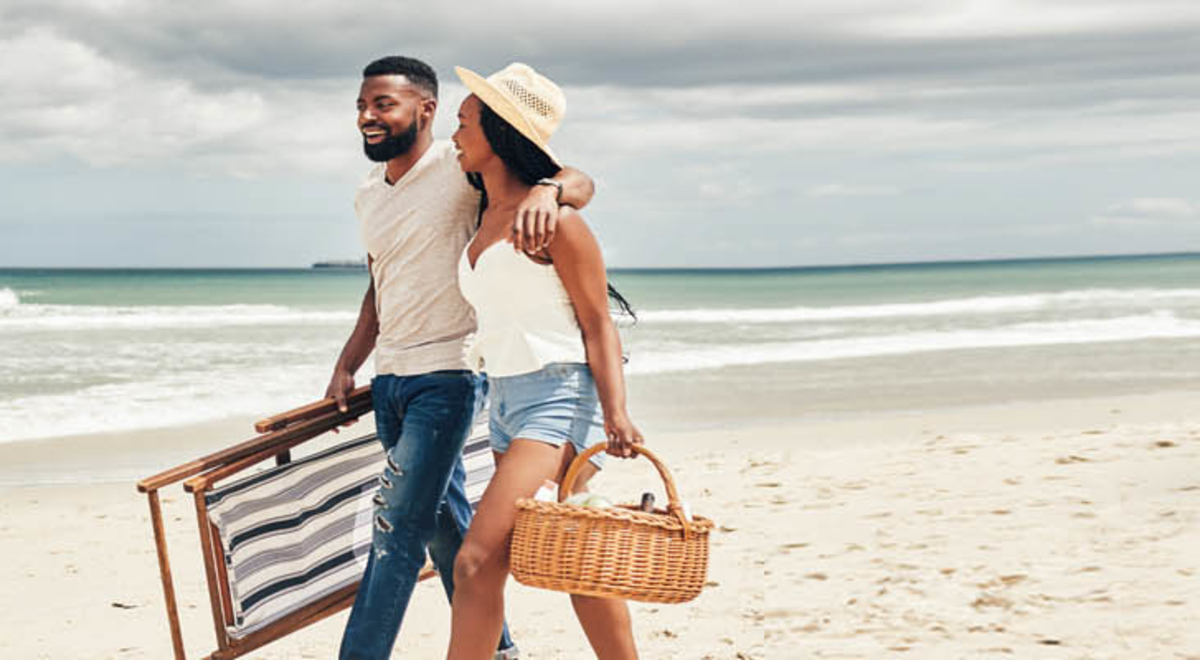 A couple hugging and walking on the beach as they hold a chair and a picnic basket