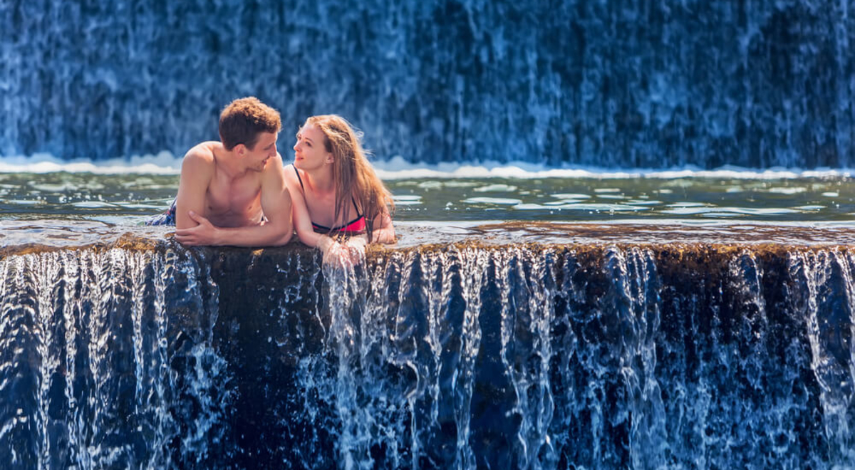 A couple swim to the edge of a giant pool between two waterfalls