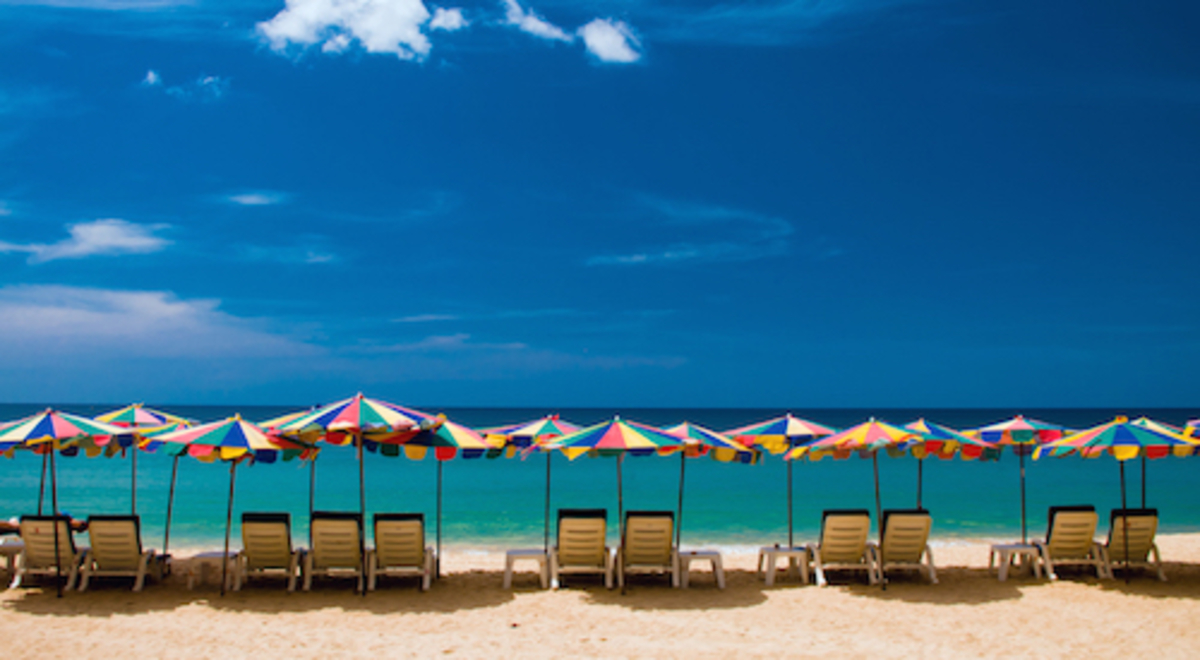 Deck chairs with colourful umbrellas line a golden-sanded beach with a still blue sea and sky in the background.