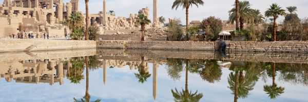 Egyptian obelisks and sandstone ruins lined by palm trees and a clear lake