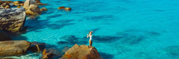 Drone shot of a lady standing on a large rock in shallow, blue water
