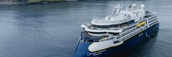 An aerial view of the National Geographic Endurance expedition cruise ship, with its distinctive navy blue hull and yellow trim, sailing on dark calm water with dramatic green and brown mountain ranges rising under an overcast sky in the background.