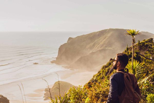 Bearded man wearing a backpack staring out at the ocean from atop a grassy cliff