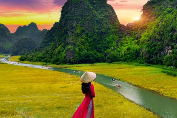 Woman in a red Chinese dress looking out at green fields, a river, and tall, forested mountains at sunset