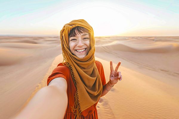 Person with scarf around head smiling and holding up peace sign while standing in the desert