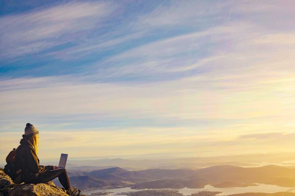 Wide shot of a person using a laptop sitting on a hill overlooking a lake-side city at sunset