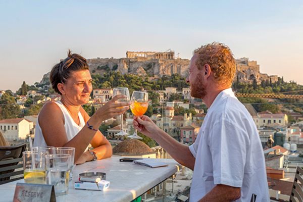 couple sitting on top of building cheersing drinks over a table with athens in the background