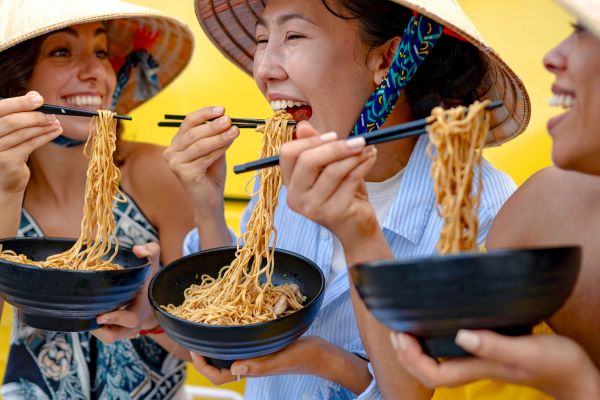 Three people smiling, eating noodles with chopsticks, wearing straw hats