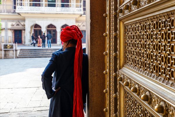 Man wearing a red turban with his back to an ornate bronze door