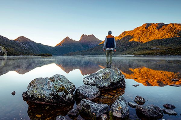 Man standing on a rock looking out at a reflective lake and tall mountains