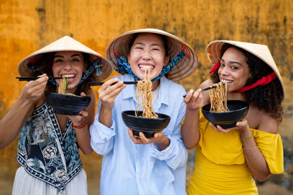 Three women enjoying noodles in front of a yellow wall