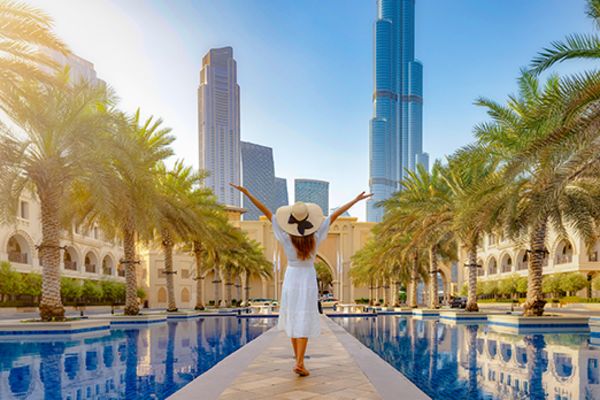 Woman in a white dress spreading her arms standing between two pools of water lined by palm trees looking up at skyscrapers